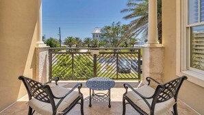 Un balcon avec une table et deux chaises, donnant sur une rue bordée de palmiers et sur un immeuble en arrière-plan.