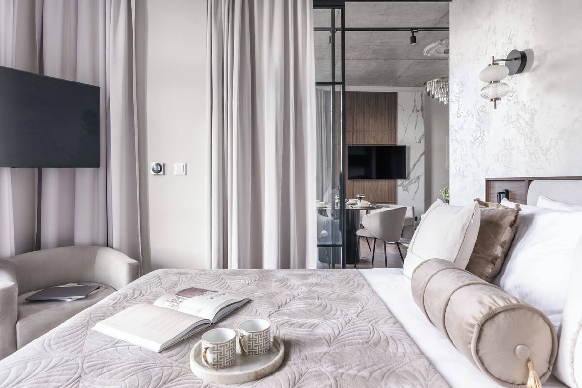 Une chambre d’hôtel moderne dotée d’un grand lit, d’un téléviseur à écran plat, d’un bureau et d’un coin repas, le tout visible à travers une paroi en verre.