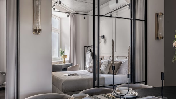Une chambre d’hôtel moderne dotée d’un grand lit, d’un coin repas avec une table dressée pour deux personnes, et offrant une vue sur une autre chambre à travers une paroi en verre.