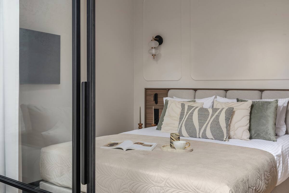 Un lit bien fait, recouvert d’une courtepointe, une table de chevet avec un livre et une tasse, et un luminaire fixé au mur.