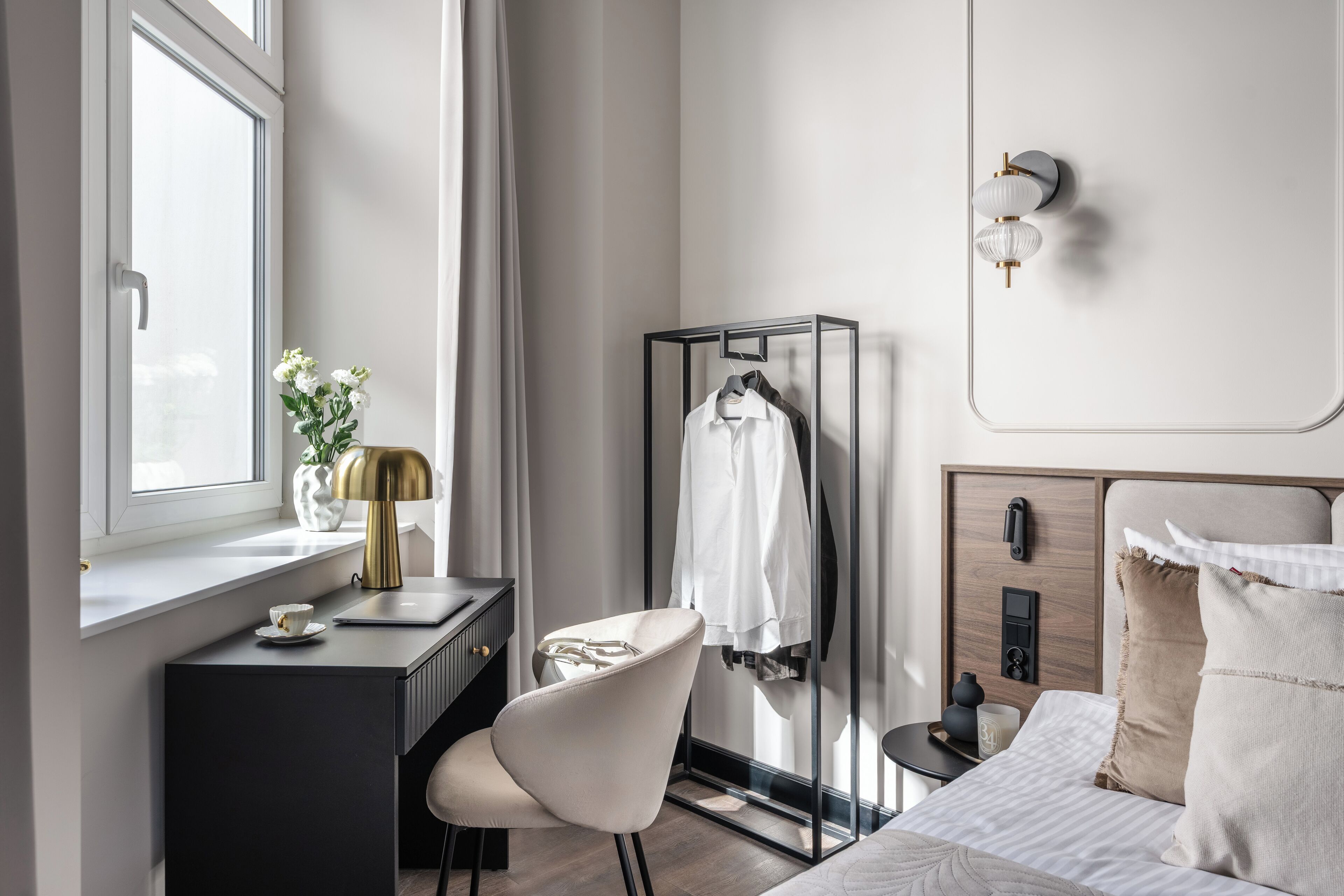 Una habitación de hotel moderna con escritorio, silla y cama con cabecera. Se observa un armario con ropa colgada, una mesita de noche con lámpara y una ventana con cortinas.