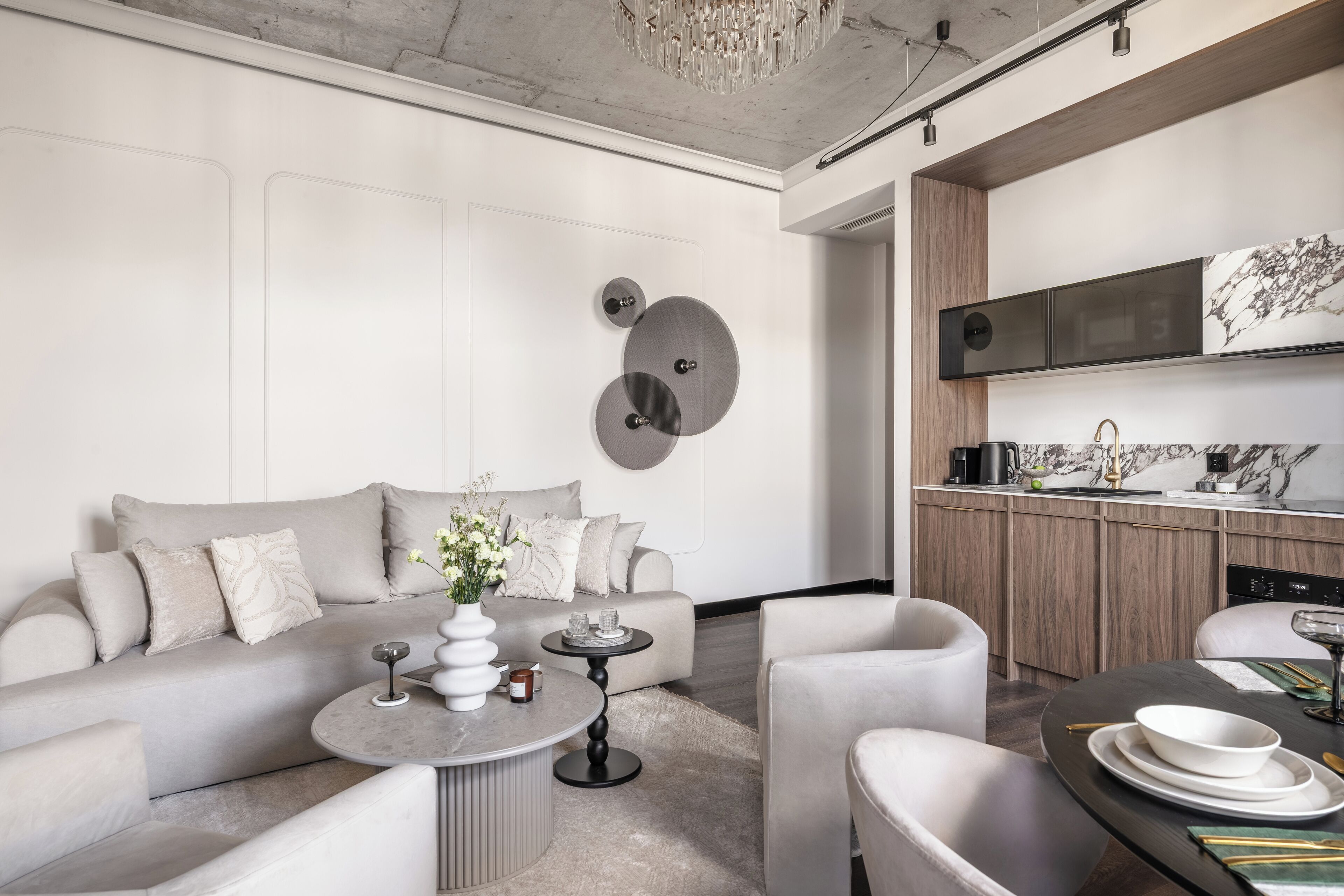 Una sala de estar moderna con sofá, mesa de centro y sillones, además de una cocina con encimera y electrodomésticos, y una araña.