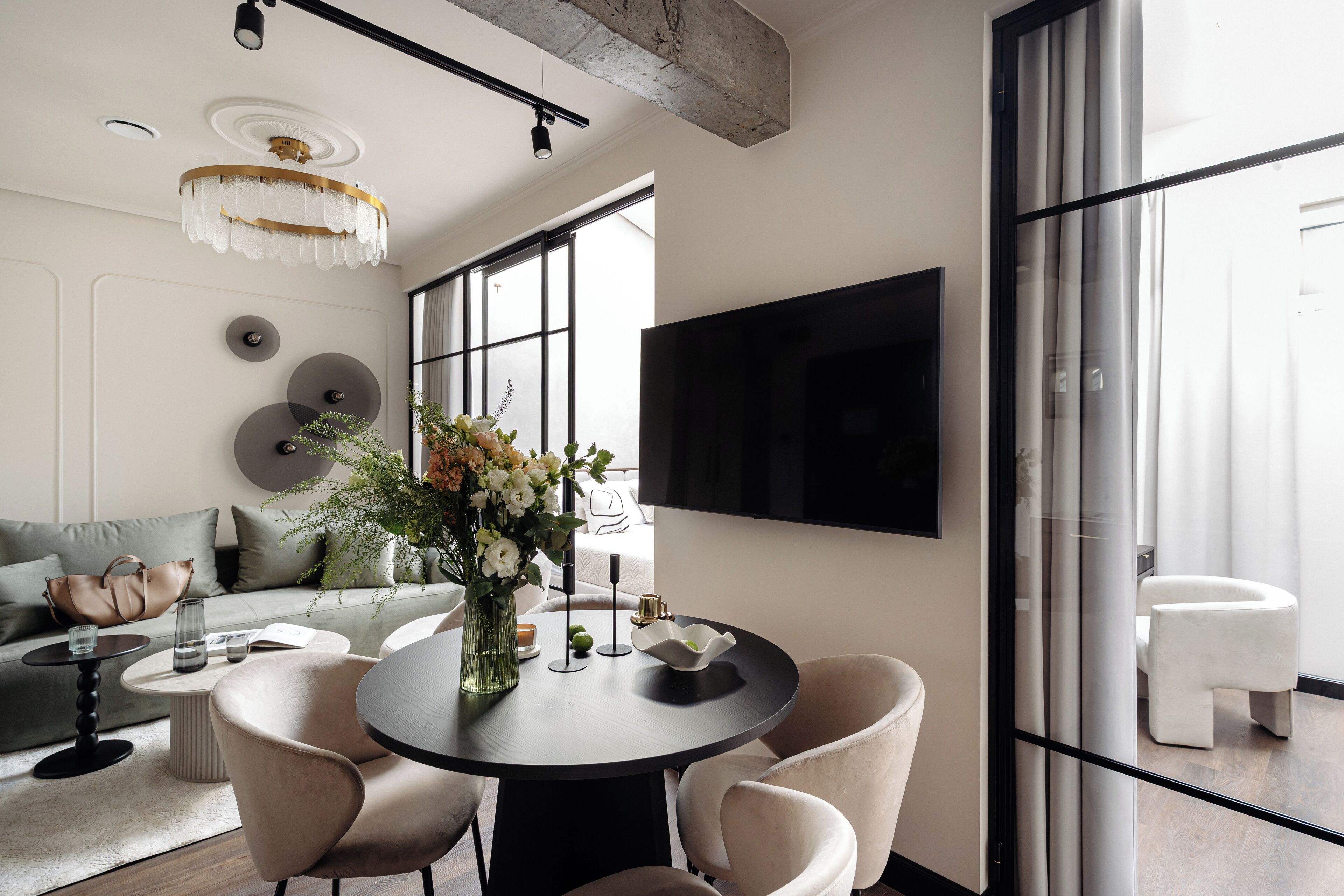 Una sala de estar moderna con una mesa de comedor redonda, asientos cómodos, un ventanal grande y un televisor montado.