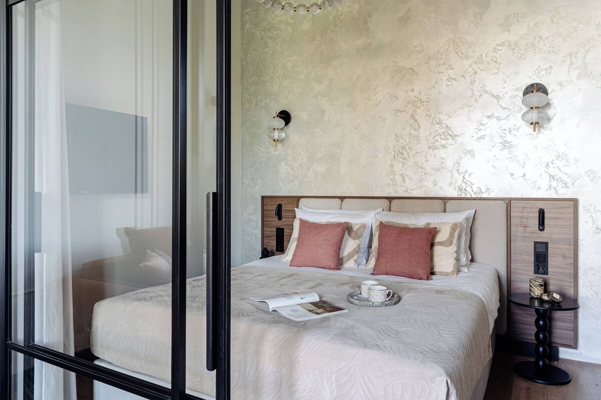 Une chambre d’hôtel moderne avec un grand lit, une table de chevet, un luminaire fixé au mur et une vue à travers une porte vitrée.