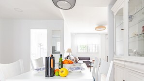 Et lyst spiseområde med et hvidt bord dækket til to, en skål med frugt og en vinflaske.