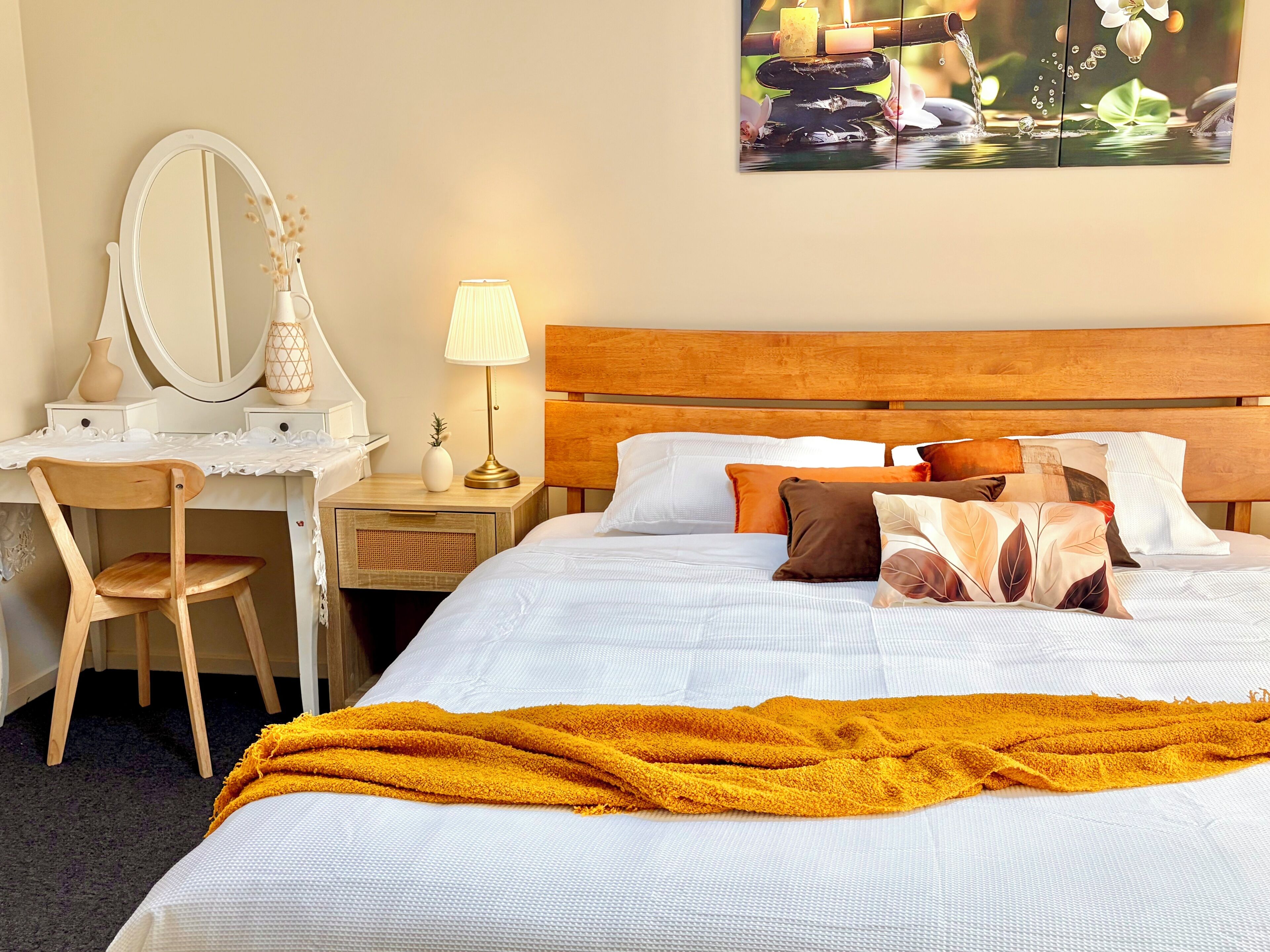 A hotel room with a wooden headboard, a bed with white bedding, a desk with a mirror, a lamp, and a vase with dried flowers.