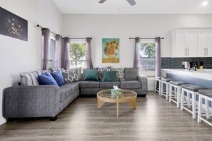 A modern living room with a grey sectional sofa, a wooden coffee table, and a kitchen area with white cabinets and a bar stool.
