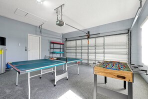 A room with a ping pong table, a foosball table, a ceiling-mounted projector, a white door, and a garage door.