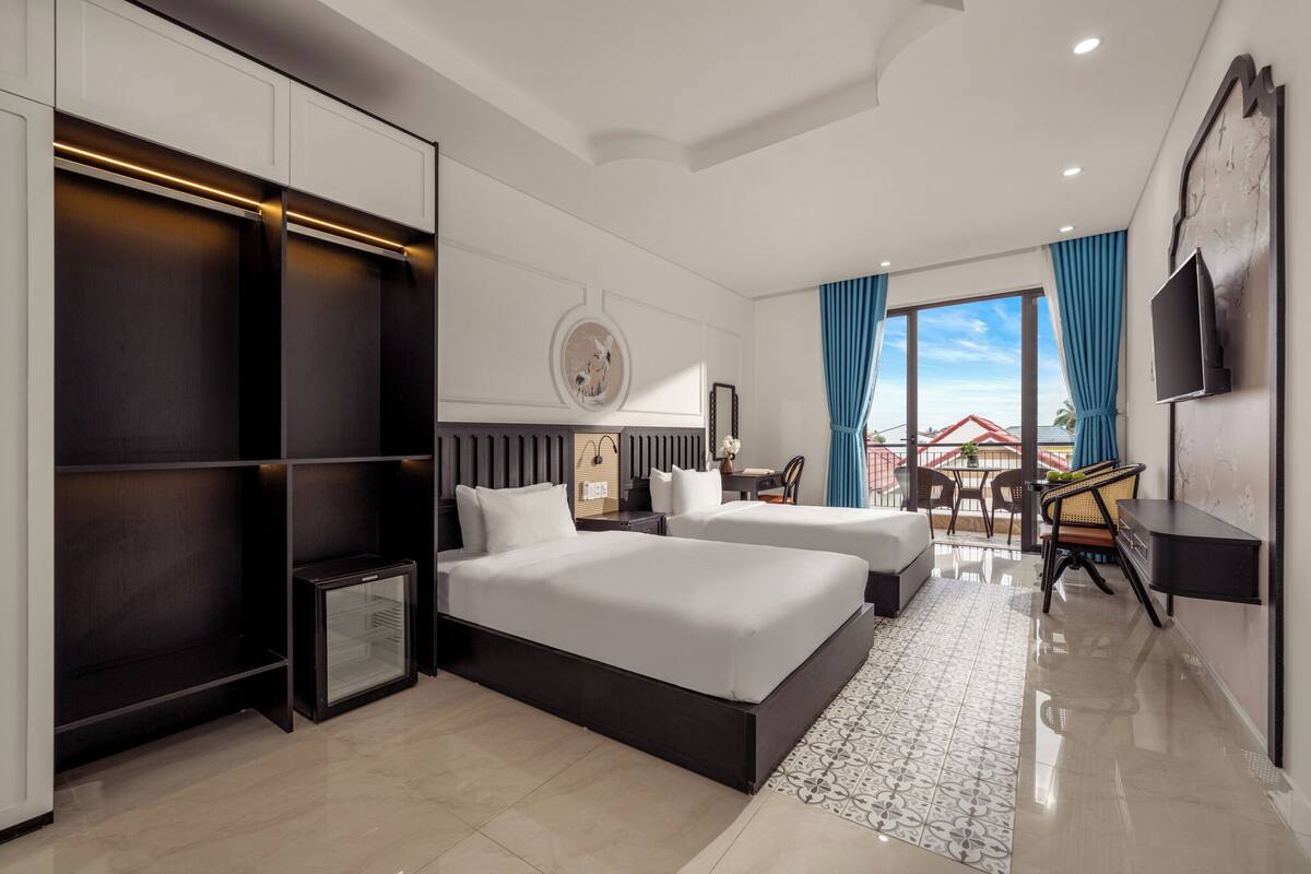 Una habitación de hotel moderna con una cama grande, un escritorio, una silla, un refrigerador pequeño y un balcón con cortinas azules.