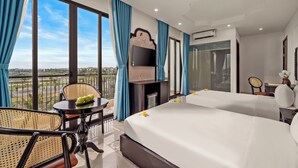 Une chambre d’hôtel avec deux lits, une télévision, un balcon avec des rideaux bleus et une table sur laquelle se trouve un bol vert.