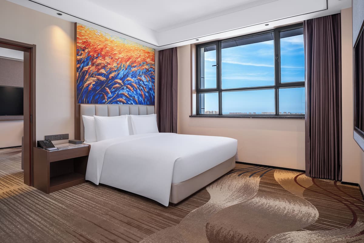 A hotel room with a large bed, a desk with a telephone, a painting on the wall, and a large window with a city view.