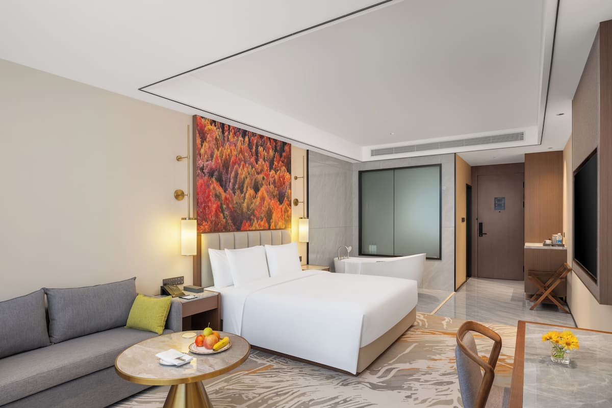 A modern hotel room with a bed, a sofa, a round table with fruit, a painting, and a bathroom with a bathtub and mirror.