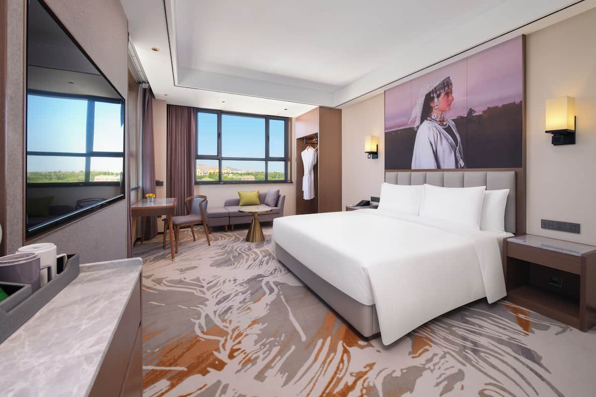 A modern hotel room with a large bed, a seating area with a sofa and chairs, a desk with a chair, and a large window offering a view.