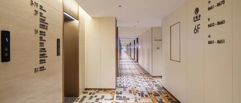 A hotel corridor with floor-to-ceiling walls, numbered room signs, and a patterned floor.