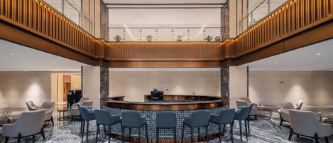 A modern hotel lobby with a central bar, seating area, and a mezzanine level.