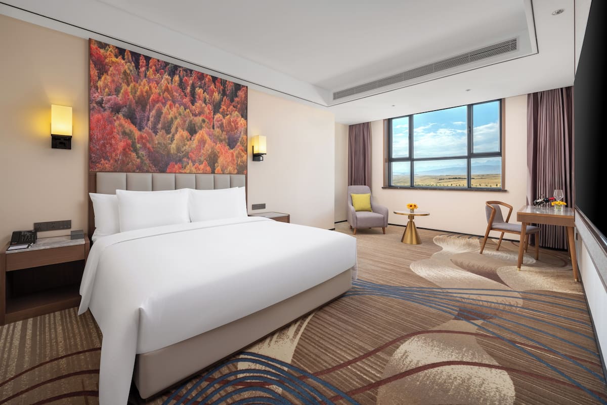 A hotel room with a large bed, a desk, a chair, a view of the outdoors, and a painting on the wall.