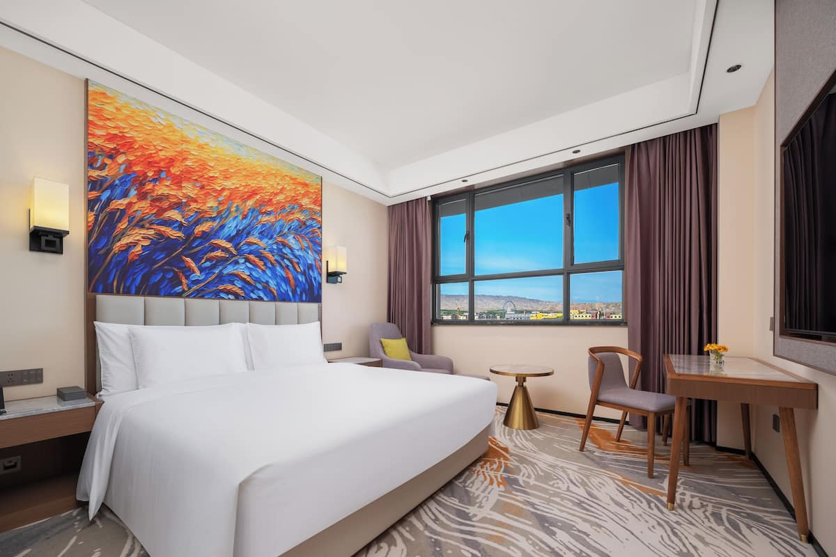 A hotel room with a large bed, a desk, a chair, a small table, a painting on the wall, and a window with a view of a cityscape.
