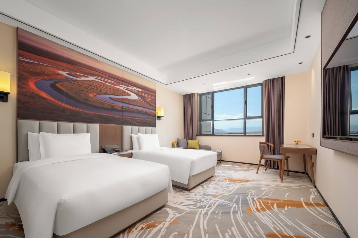 A modern hotel room with two beds, a large abstract painting on the wall, a desk with a chair, and a large window offering a view.