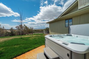 Relax in the hot tub on the patio, surrounded by a grassy lawn and trees.