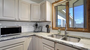 A kitchen with white cabinets, a granite countertop, a stainless steel sink, a microwave, and a window with a wooden frame. - Sunset Ridge (Penrose)
