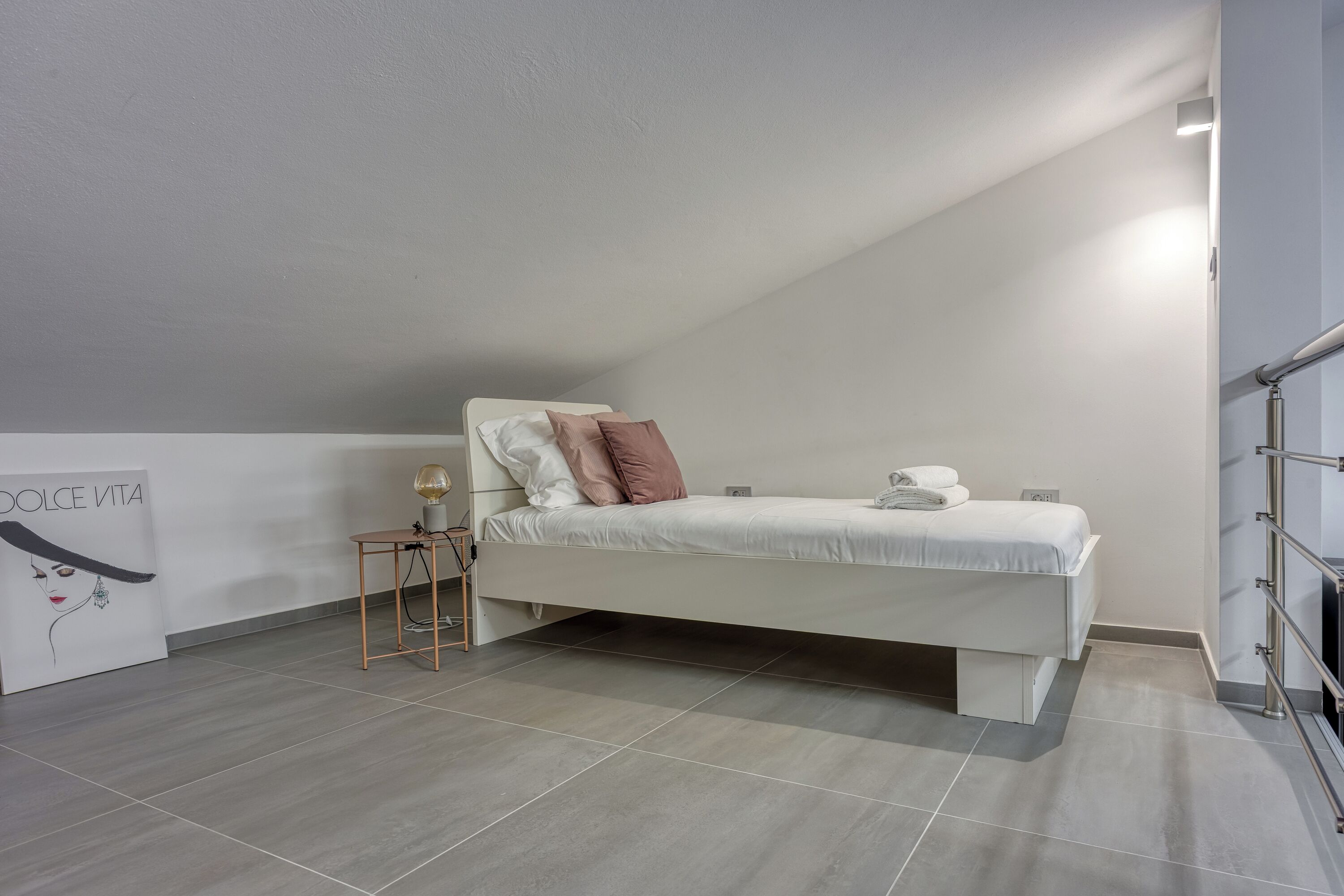 Ein modernes Schlafzimmer mit einem weißen Bett, einem kleinen Nachttisch, einer Lampe und einem wandmontierten Regal.
