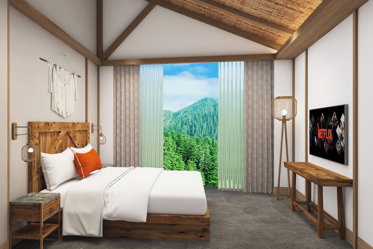 Ein Schlafzimmer mit Holz-Kopfteil, einem Bett mit weißer und orangefarbener Bettwäsche, einem Nachttisch, einer Wandlampe, einem großen Fenster mit Blick auf die Berge und einem an der Wand befestigten Fernseher, der das Netflix-Logo zeigt.