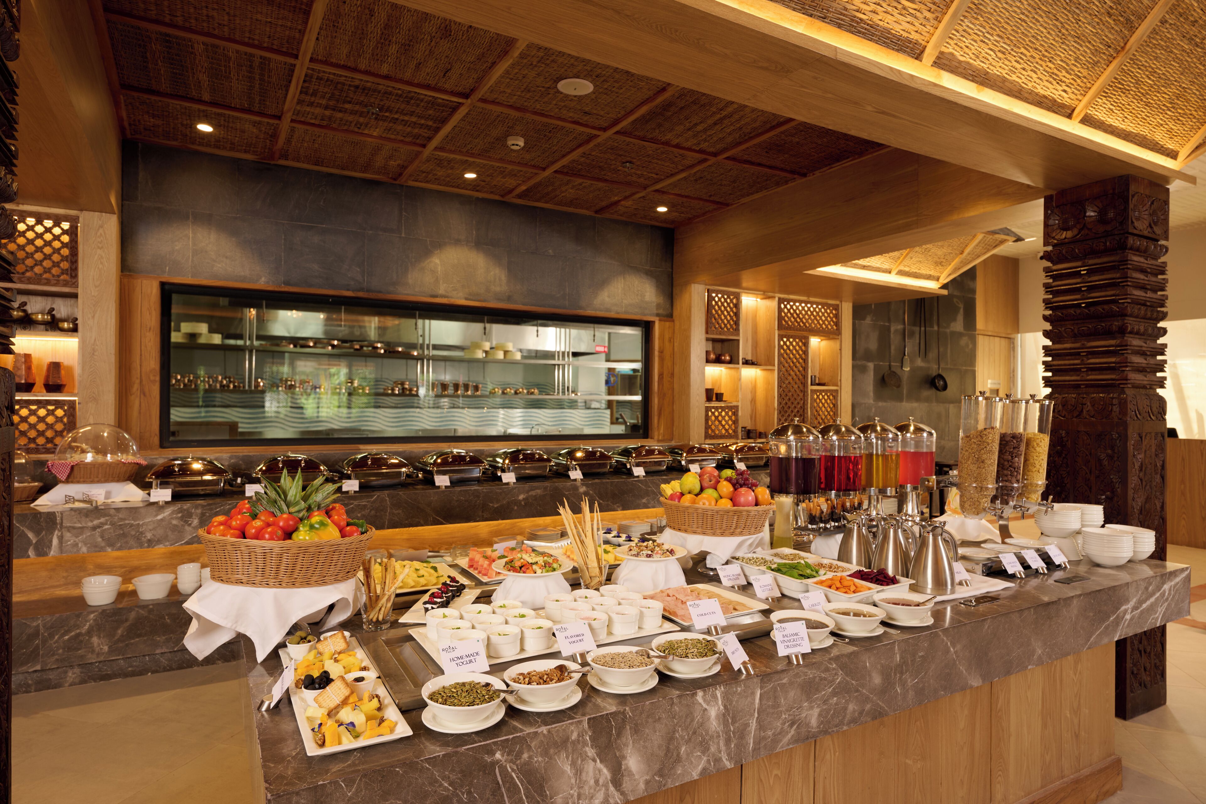 Un buffet proposant une variété de plats et de boissons, un coin cuisine à l’arrière-plan et un plafond en bois orné d’un motif tissé.