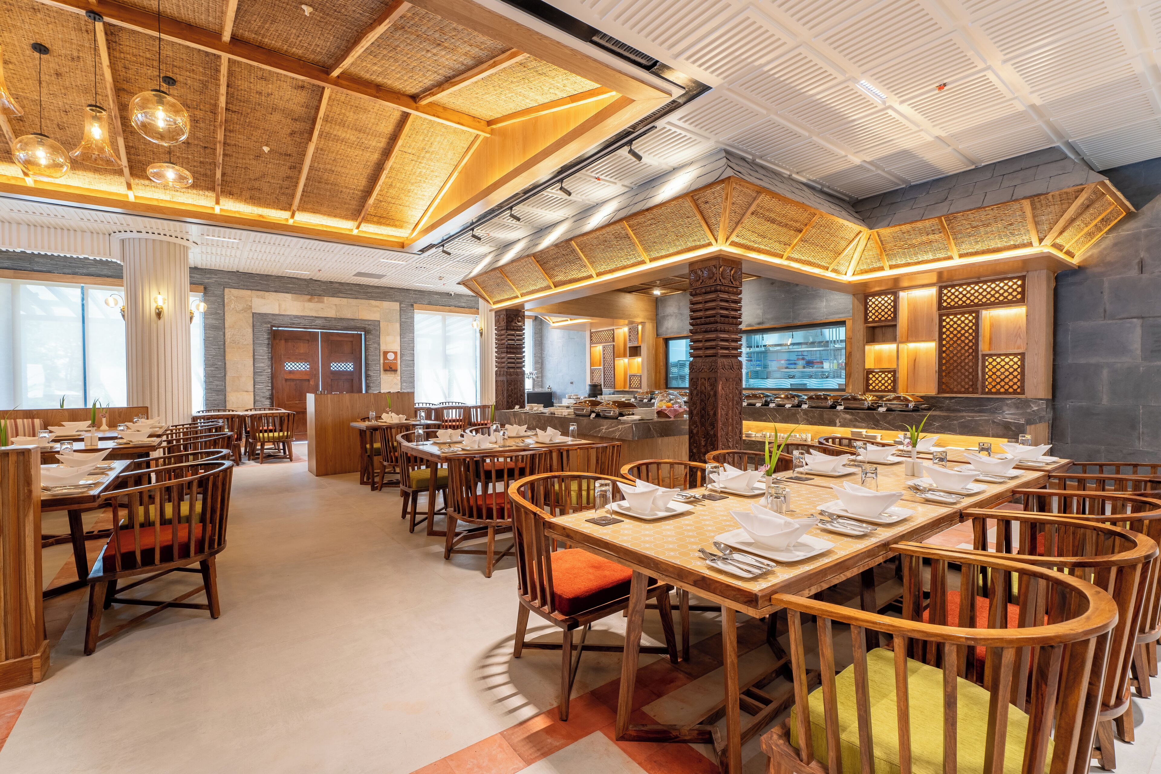 Ein geräumiges Restaurant mit Holztischen und -stühlen, einer hohen Decke mit sichtbaren Holzbalken und einer modernen Küche im Hintergrund.