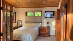 A bedroom with a bed, a dresser, a mirror, a window with blinds, and a television on the wall. - Auke Bay View Studio (Juneau)