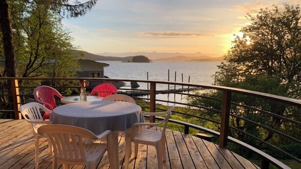 A wooden deck with a round table and chairs, overlooking a body of water and distant mountains. - Auke Bay View Studio (Juneau)