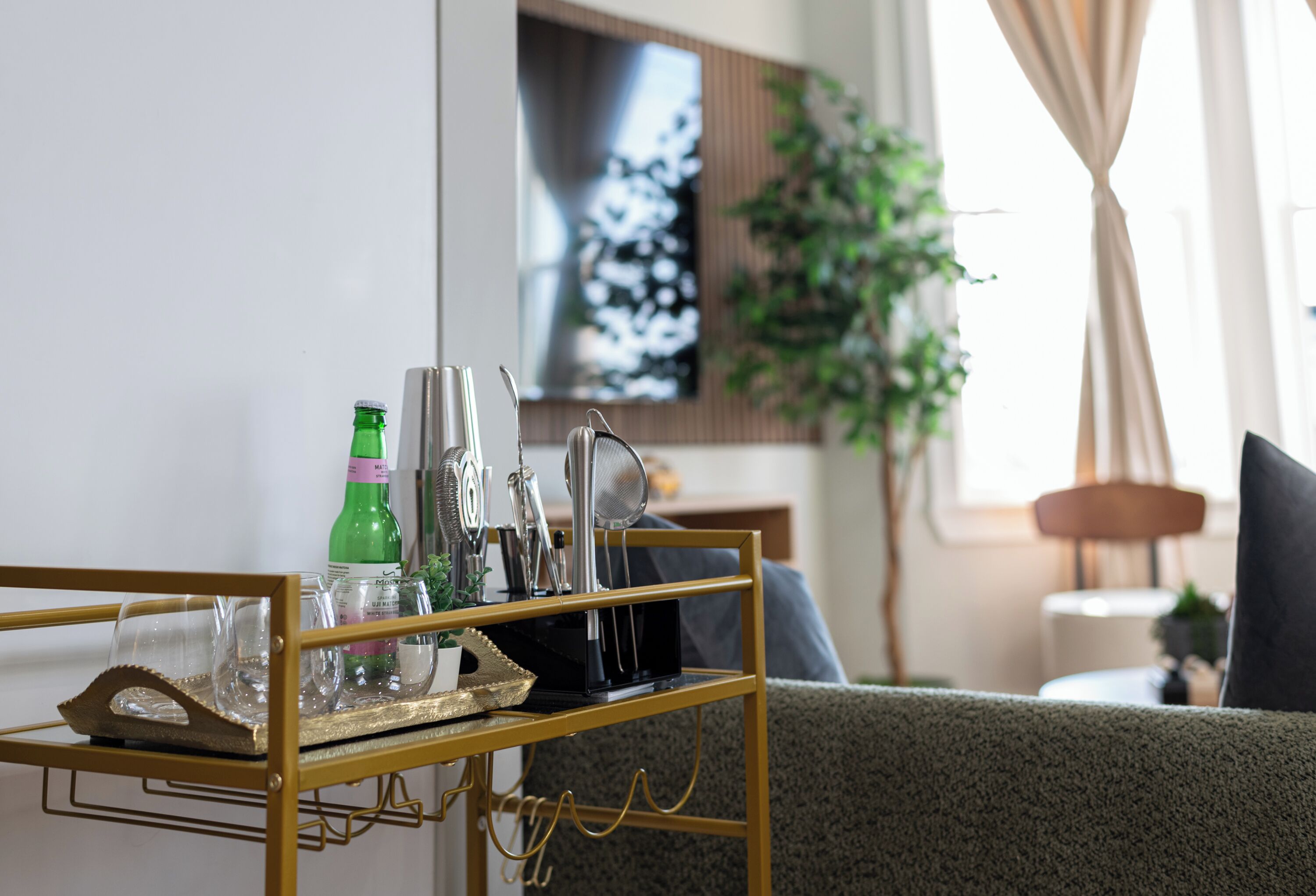 Un salon moderne avec une table d’appoint à cadre doré sur laquelle sont posés des verres, une bouteille verte et une machine à café. Un téléviseur à écran plat est fixé au mur, et une plante est visible à l’arrière-plan.