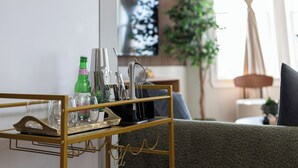A modern living room with a gold-framed side table holding glasses, a green bottle, and a coffee maker. A flat-screen TV is mounted on the wall, and a plant is visible in the background. - Cozy 3BR Stylish Home near Bus+PATH Access to NYC Metlife with Skylight Family (Jersey City)