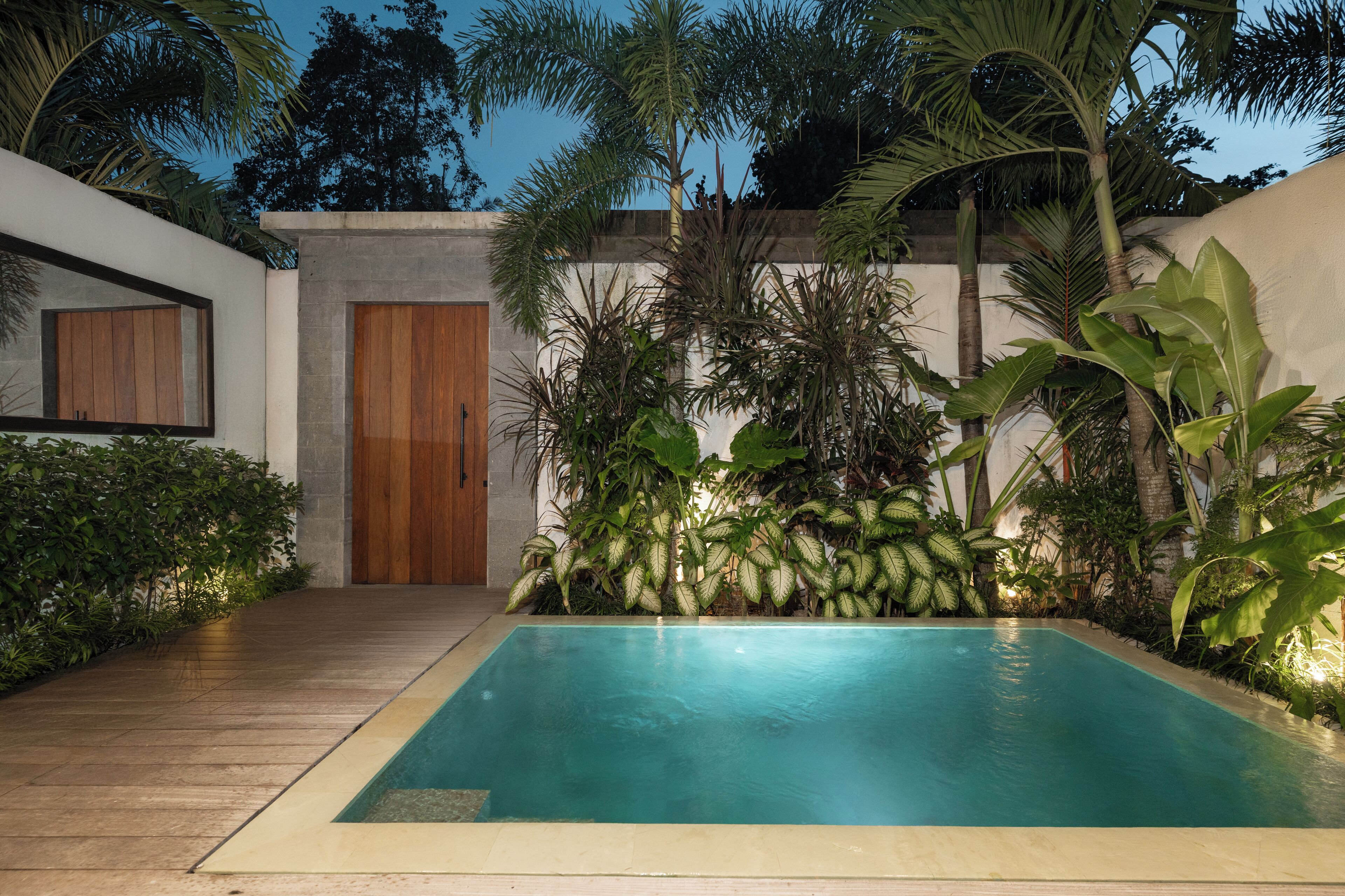 A rectangular pool with a wooden deck surrounding it, set against a backdrop of tropical plants and a modern building with a wooden door.