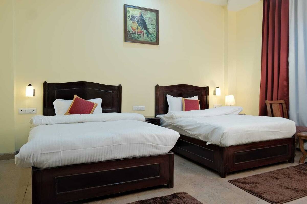 Habitación de hotel con dos camas individuales, cada una con cabecero de madera, ropa de cama blanca y almohadas rojas decorativas. Hay un cuadro en la pared sobre las camas y una cortina roja en la ventana.