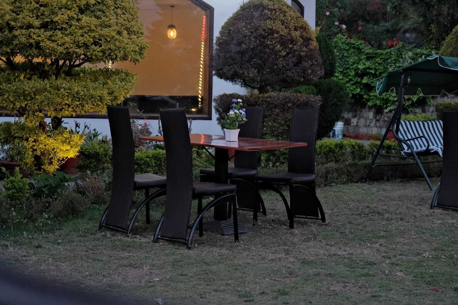 Un espace de restauration en plein air avec une table et des chaises, entouré de verdure et bordé d’un bâtiment dont la surface réfléchit la lumière.