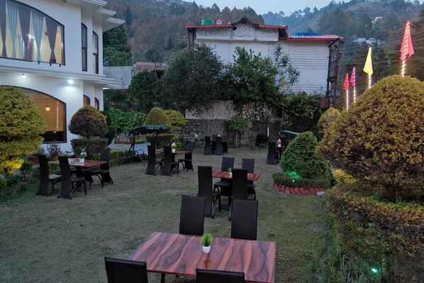 Un espacio de comedor al aire libre con varias mesas y sillas, rodeado de setos y árboles bien cuidados.