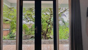 A view through a glass door to an outdoor area with a brick wall, a tiled patio, and a large tree. - Villa Kamila Senggigi By Villaloka (Senggigi)