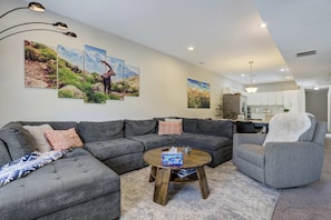 Een moderne woonkamer met een hoekbank, een ronde salontafel en een grijze fauteuil. Drie aan de muur bevestigde schilderijen tonen landschappen.
