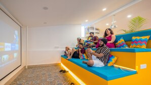 Eine Gruppe von Menschen sitzt auf einem farbenfrohen Sofa in einem Raum mit gemusterten Bodenfliesen und einer Projektionsleinwand, auf der Streaming-Dienste laufen.