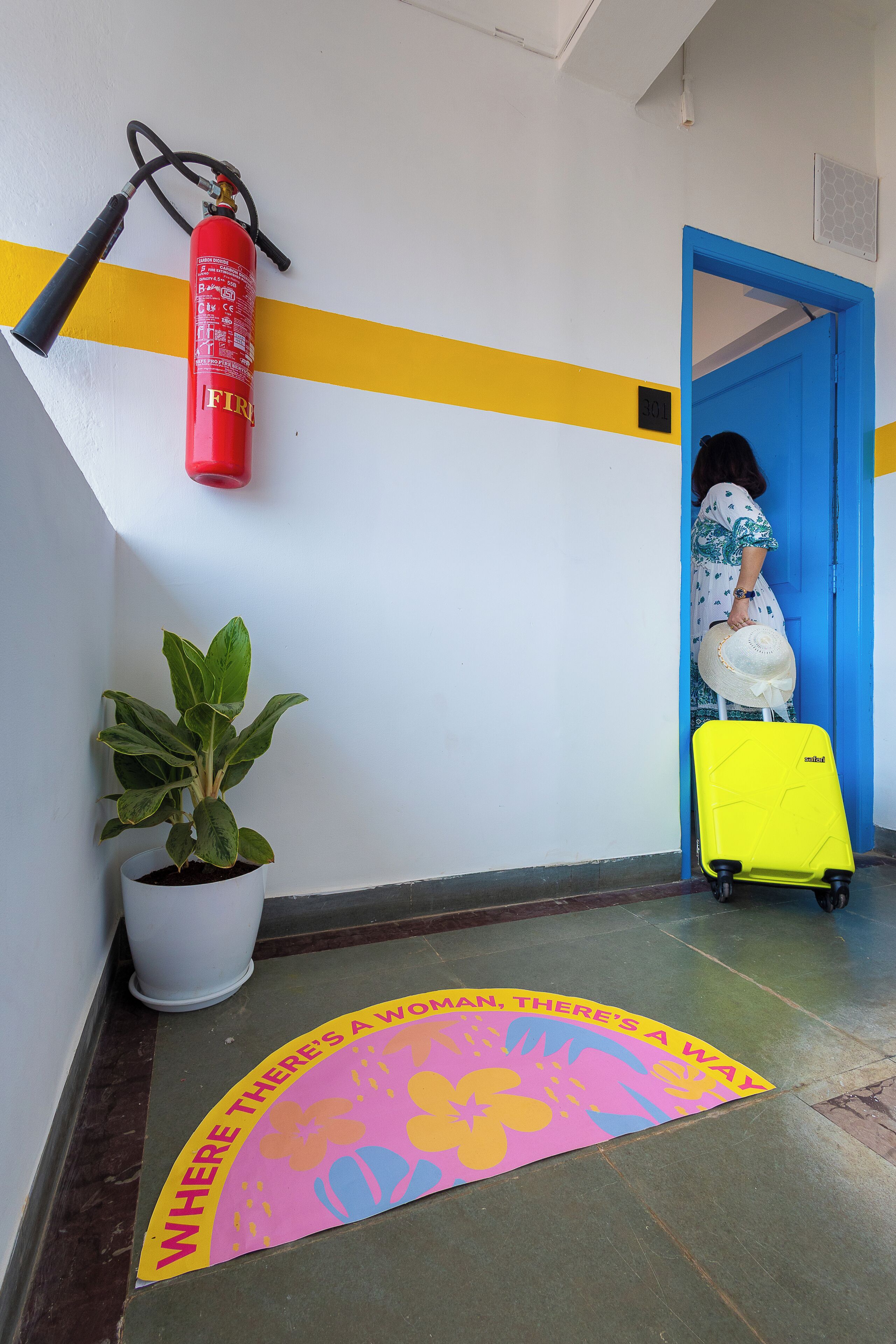 A hallway with a fire extinguisher, a potted plant, a bright yellow suitcase, and a door with a blue frame.