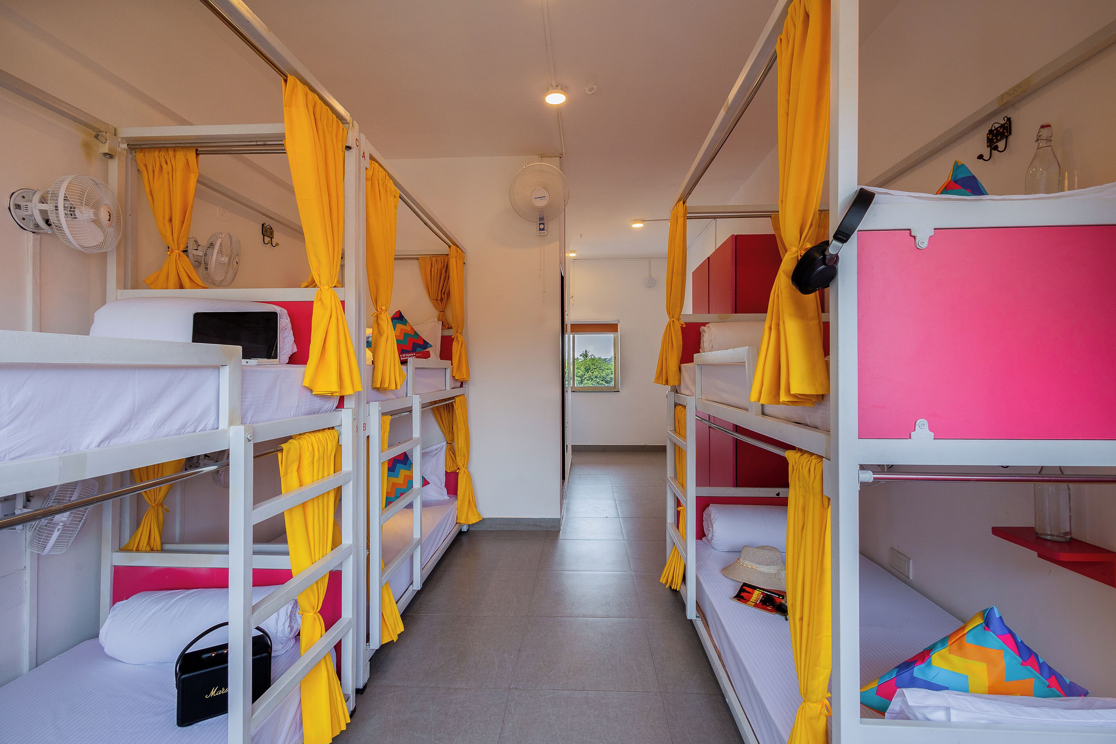 Ein Korridor mit Etagenbetten, jeweils mit gelben Vorhängen, und einem Ventilator an der linken Wand.