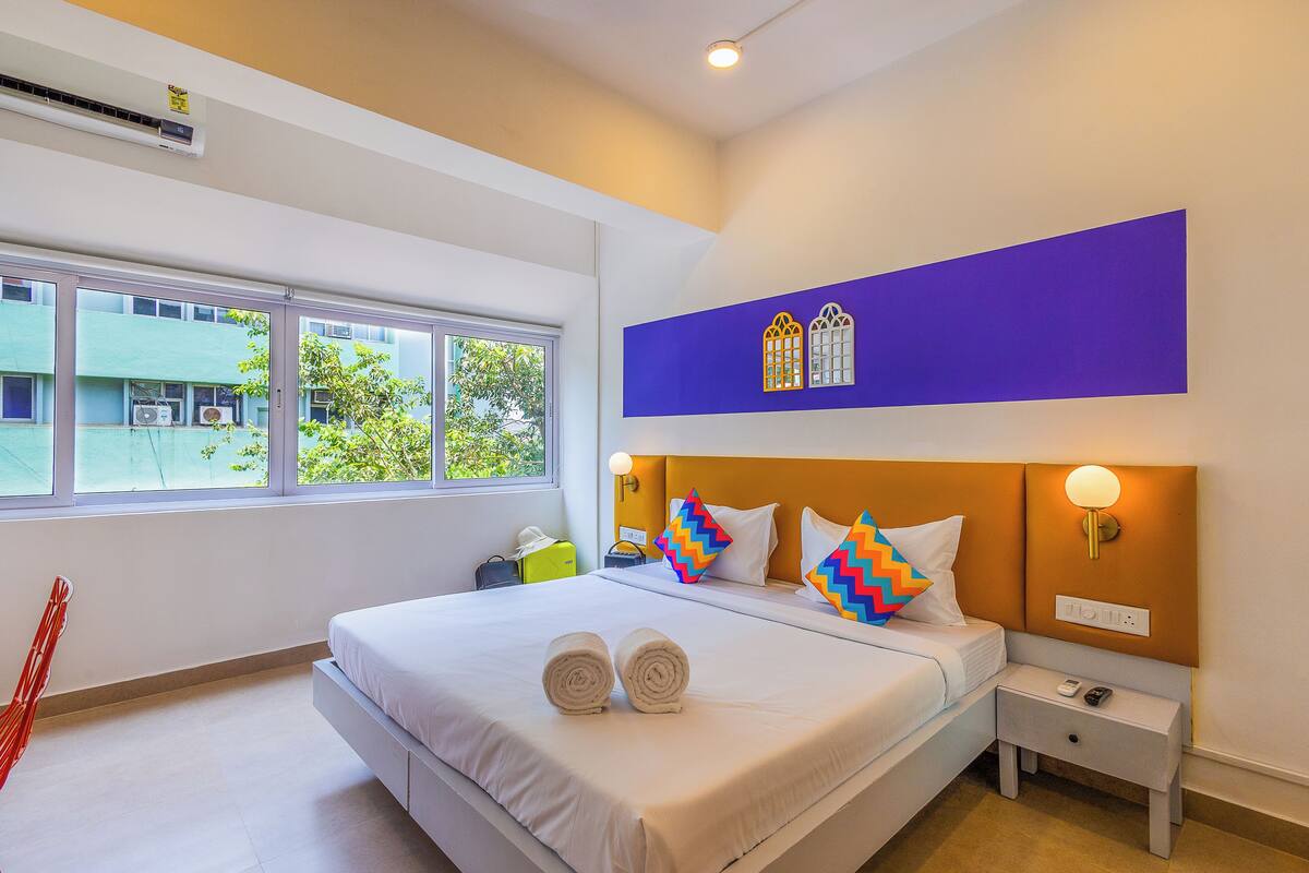 Quarto de hotel com cama, mesa de cabeceira, luminária na parede e janela com vista para edifícios e árvores.