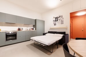 A compact room with a kitchenette, a bed, a dining table, and a wall-mounted shelf.