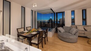 A modern living room with a dining area, large windows, and a view of the city at dusk. - Villa Blanc H (Motobu)