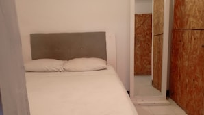 Ein kleines, ordentlich bezogenes Bett mit grauem Kopfteil in einem Zimmer mit weißen Wänden und einem Holzschrank.
