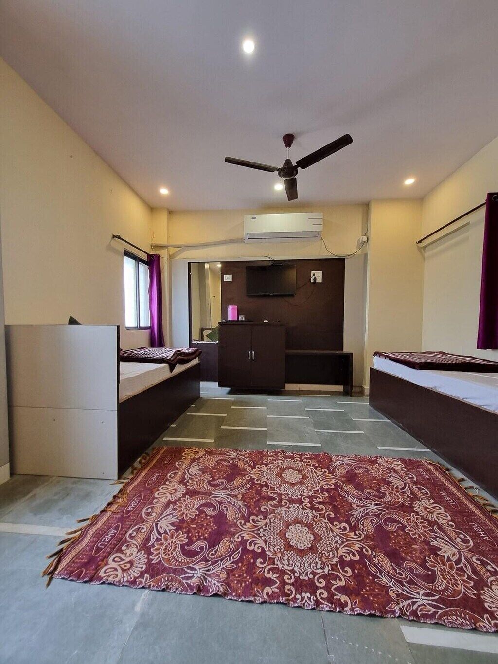 A room with a large patterned rug, a ceiling fan, an air conditioning unit, a TV, and a dark cabinet.