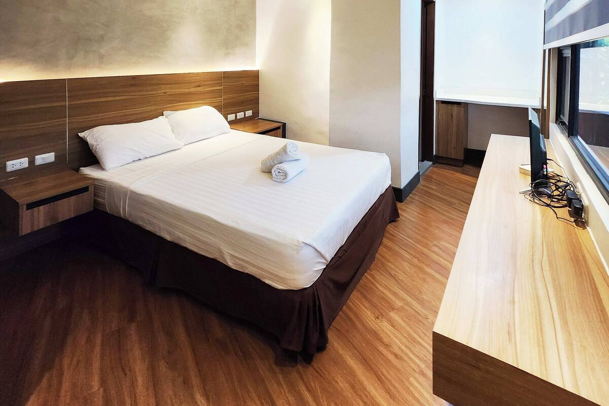 Una cama bien hecha con sábanas blancas y una toalla enrollada, una mesita de noche de madera y un escritorio de madera con una lámpara.