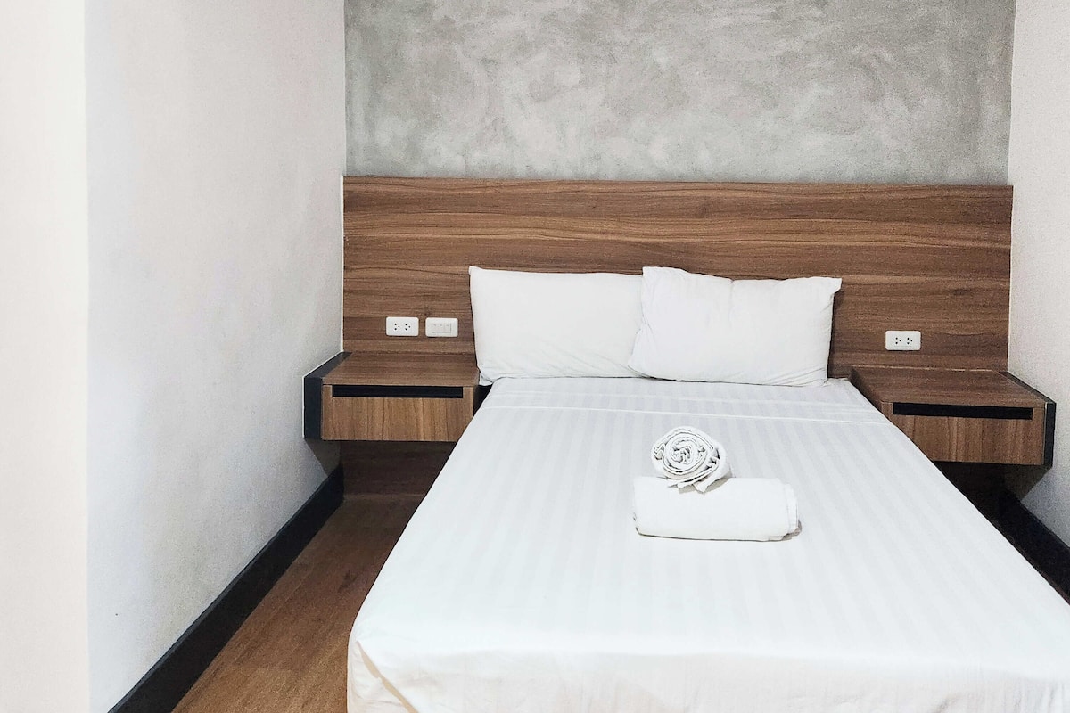 Una cama bien hecha con sábanas blancas, un cabecero de madera y una mesita de noche de madera con toma de corriente.