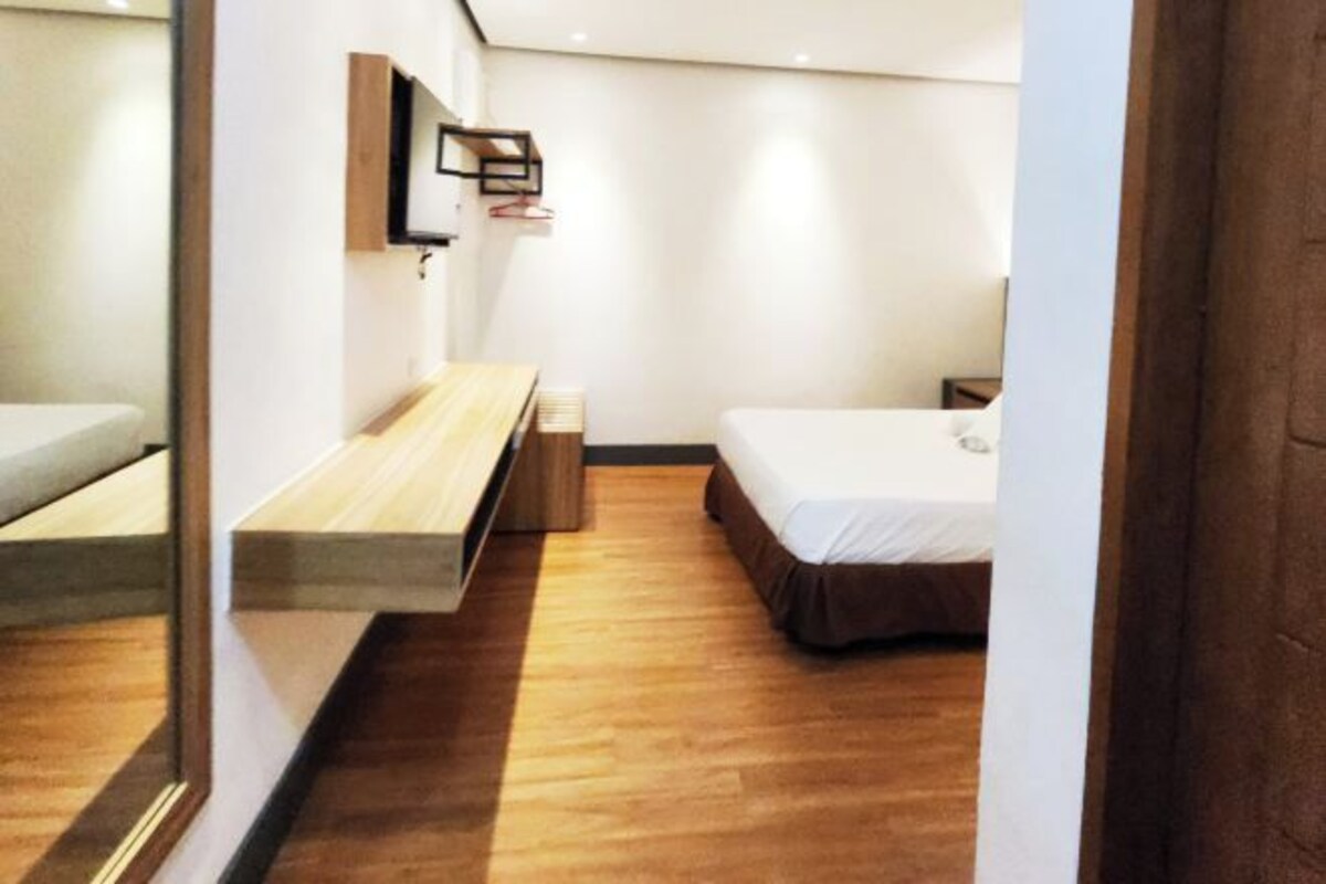 A modern hotel room with a wooden floor, a bed, a bench, a small table, and a wall-mounted TV.