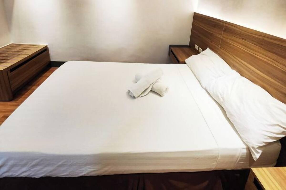 Una cama bien tendida con sábanas blancas y una toalla doblada, un cabecero de madera y una mesita de noche de madera con una bandeja.
